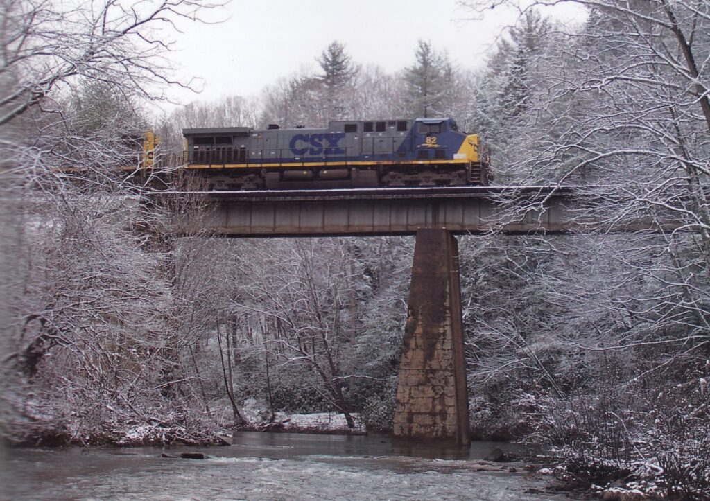 An CSX train crosses a steel bridge over a river on a snowy day.